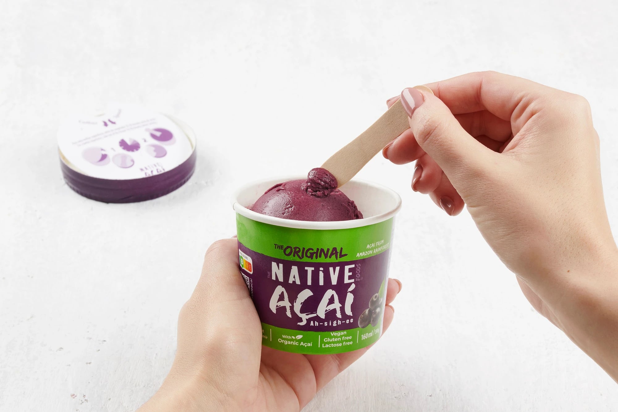Mãos seguram copo de gelado com rótulo "THE ORIGINAL NATIVE FOODS AÇAÍ" e espetam pau de madeira