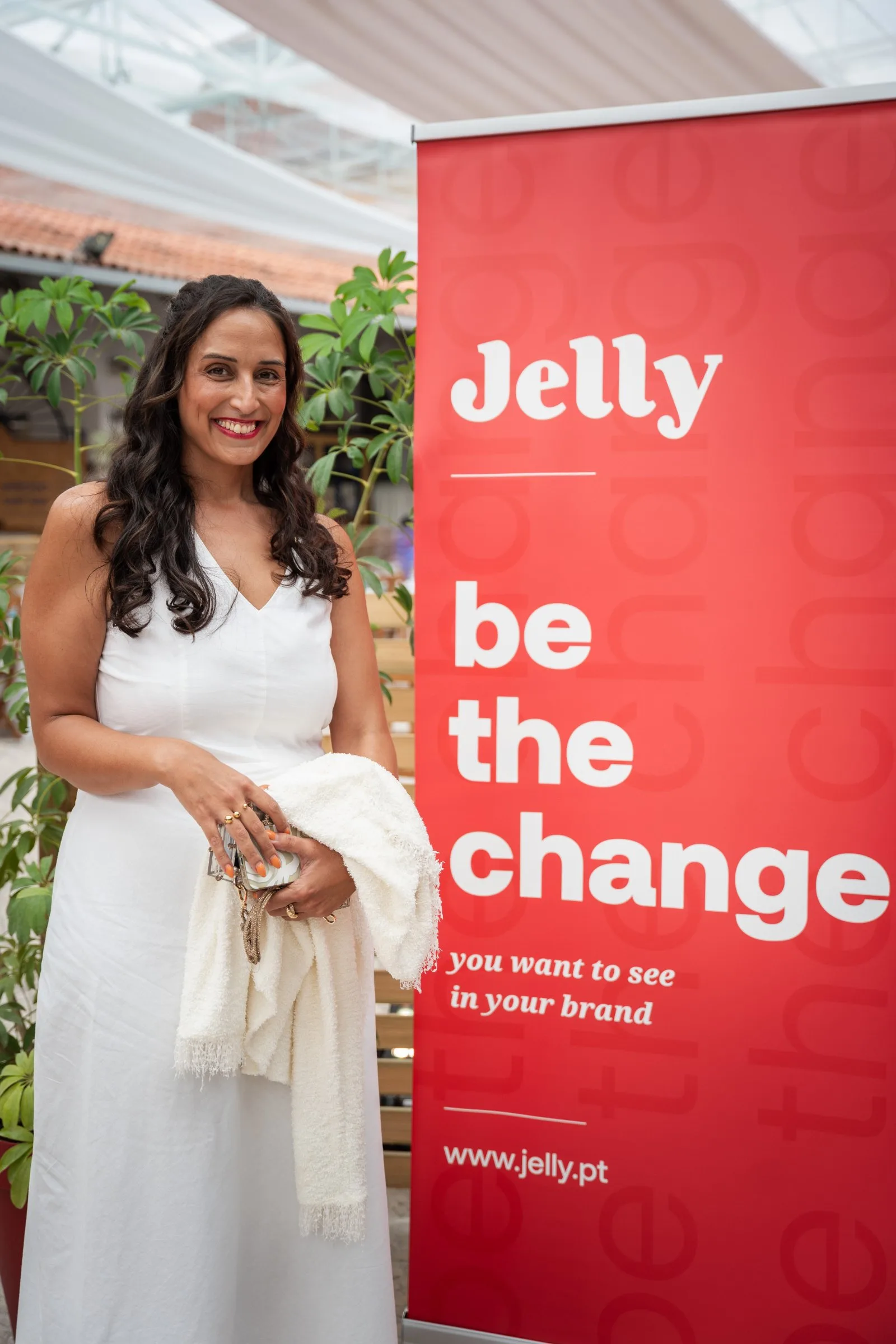 Mulher de vestido branco sorri junto a roll-up vermelho "Jelly – be the change you want to see in your brand"